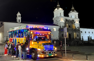 Chiva turística iluminada multicolor con grupo de personas abordando frente a iglesia colonial iluminada por la noche en Quito durante las Fiestas de la ciudad