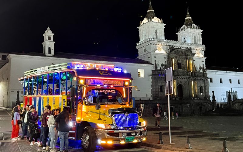 Chiva turística iluminada multicolor con grupo de personas abordando frente a iglesia colonial iluminada por la noche en Quito durante las Fiestas de la ciudad